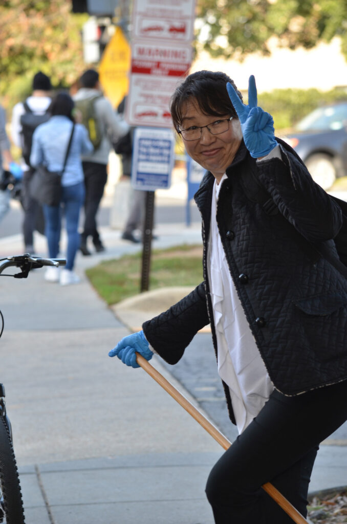 DSC_7248 Person with blue gloves giving a peace sign while holding a broom on a sidewalk, with people in the background.