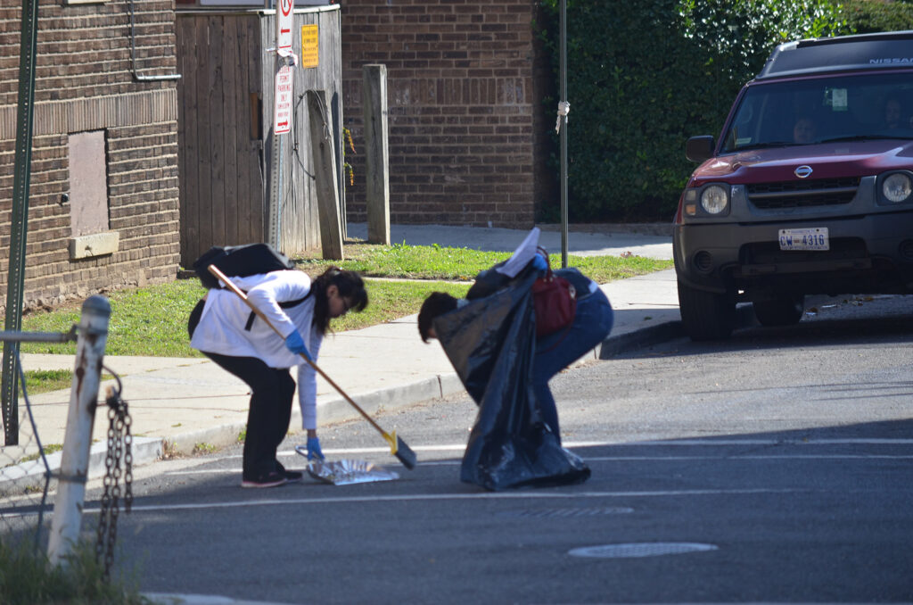 DSC_7258 Two people cleaning the street, one with a broom and dustpan, the other holding a trash bag near parked car.