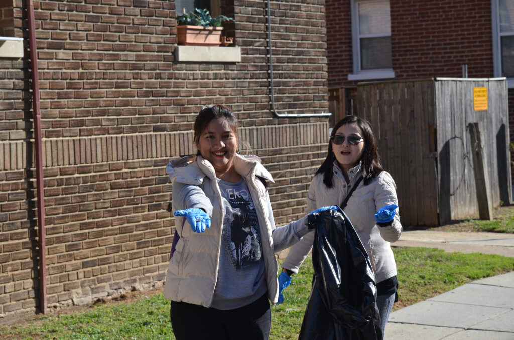 DSC_7261 Two women in jackets and gloves picking up litter outdoors on a sunny day, holding a trash bag.