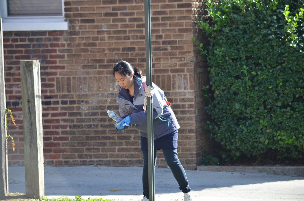 DSC_7263 A woman cleans outdoors, picking up a plastic bottle near a brick building, engaging in community cleanup efforts.