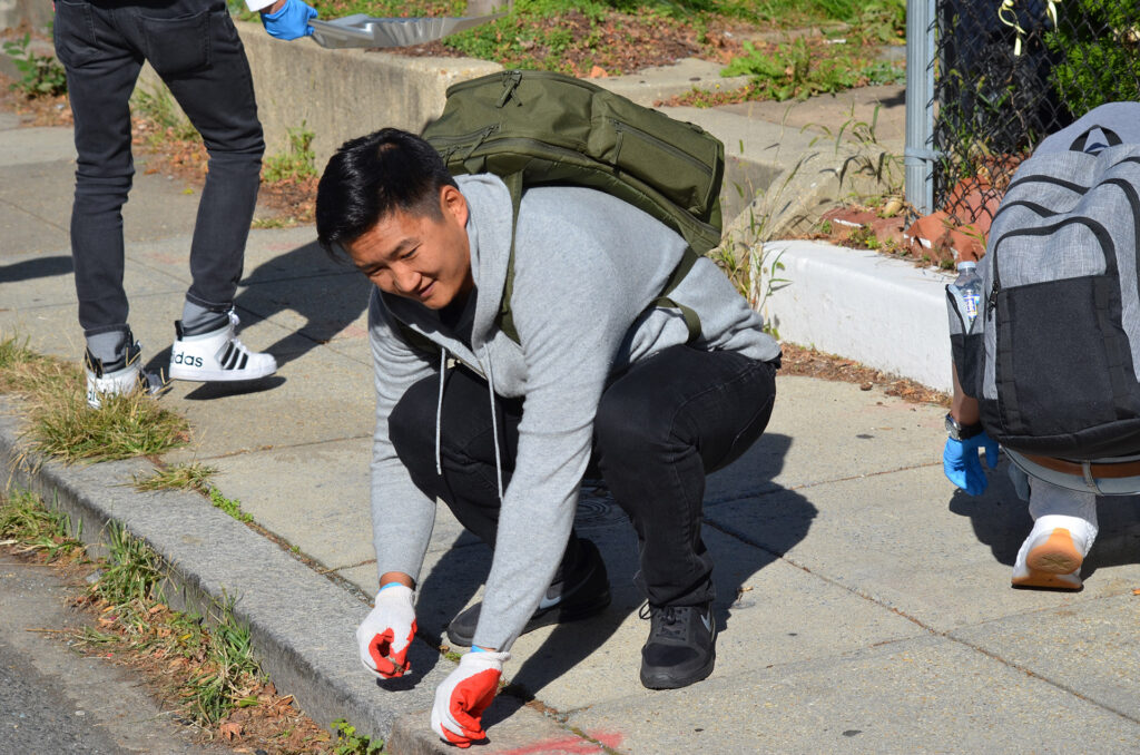 DSC_7272 Person in a gray hoodie picking up litter on a sidewalk during a community clean-up event.