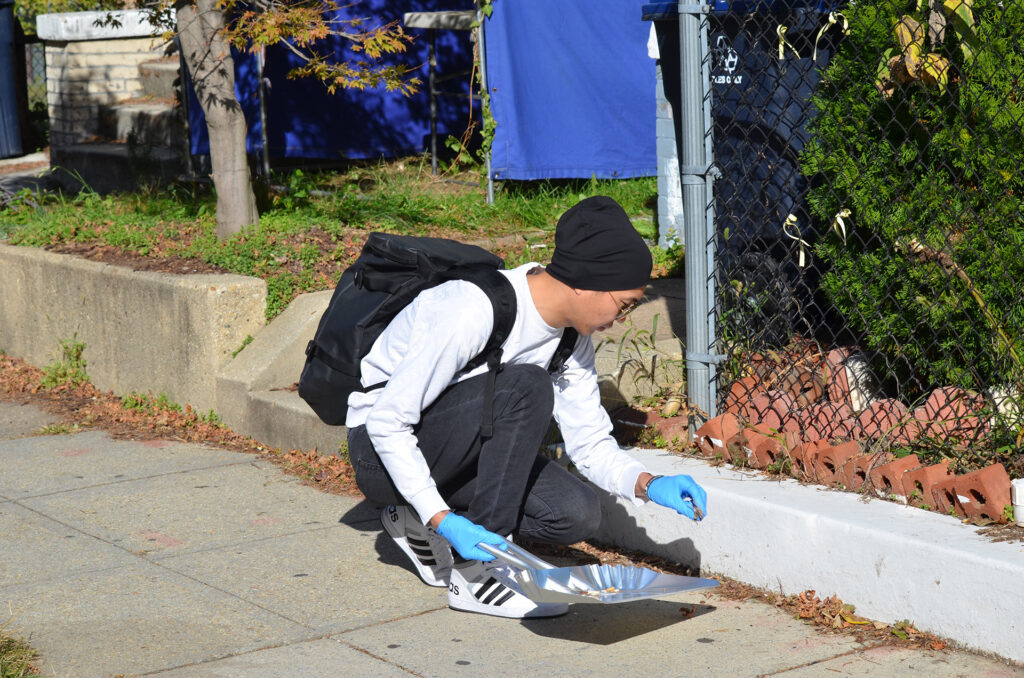 DSC_7273 A person wearing blue gloves collects leaves with a dustpan on a sidewalk near a fence.