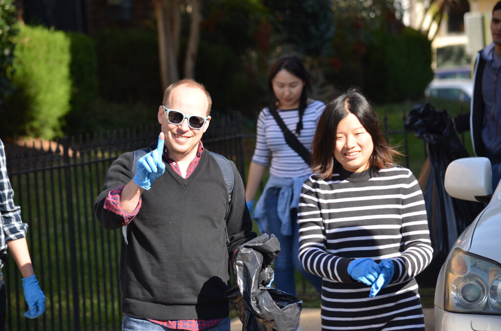 DSC_7278 People volunteering for community cleanup, smiling in sunlight while wearing gloves and holding trash bags.
