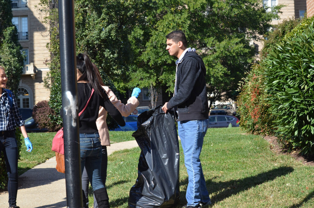 DSC_7280 People volunteering in a park cleanup, holding a trash bag and wearing gloves, with trees and buildings in the background.