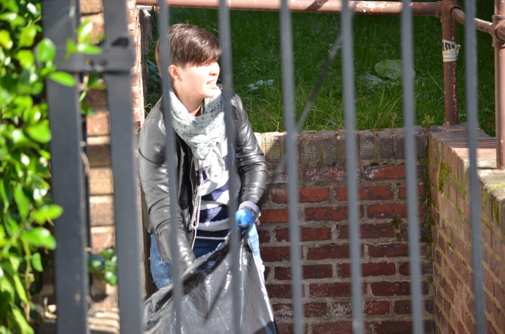 DSC_7285 Person wearing a leather jacket and scarf, cleaning behind a metal fence with gloves, urban outdoor setting.
