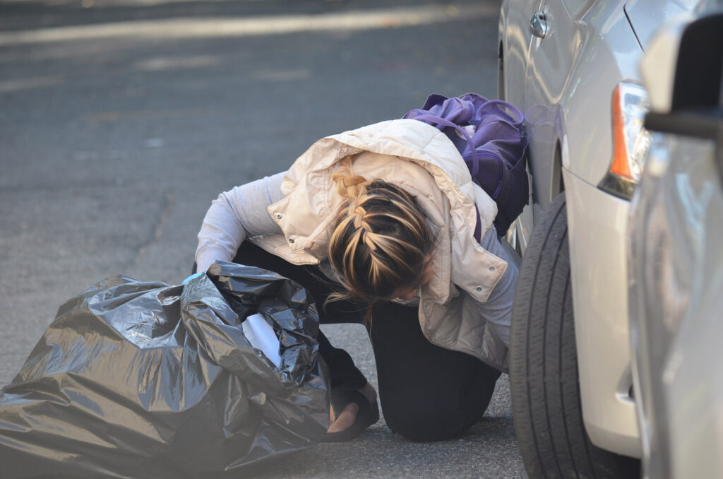 DSC_7289 Person crouching on a street, examining a trash bag beside a parked car in daylight.