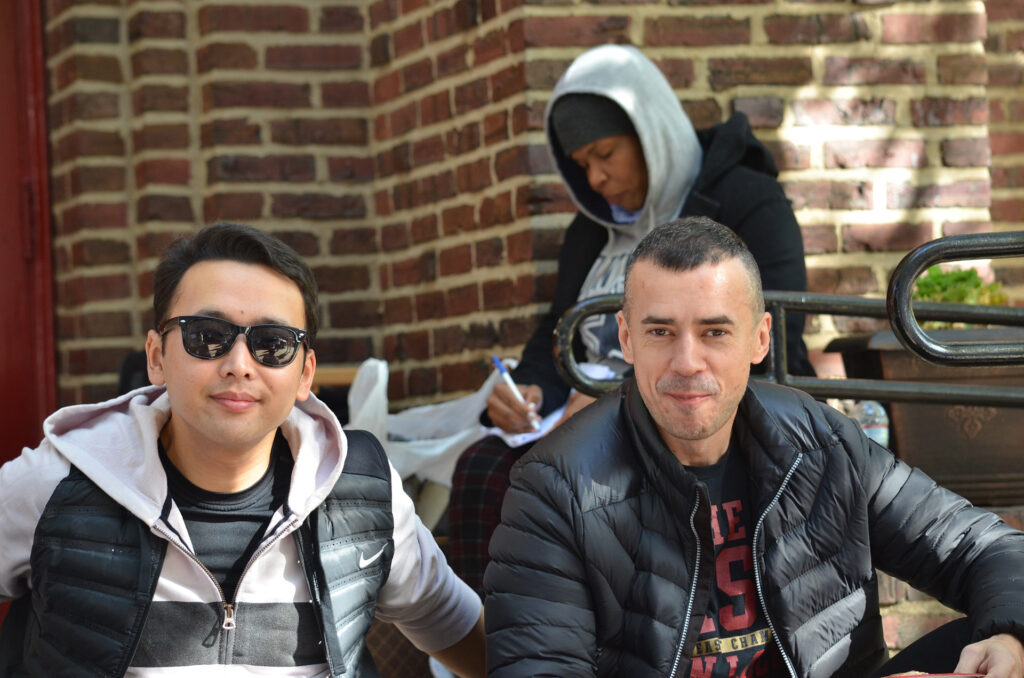 DSC_7307 Two friends smiling outdoors in winter jackets, with a person writing in the background against a brick wall.