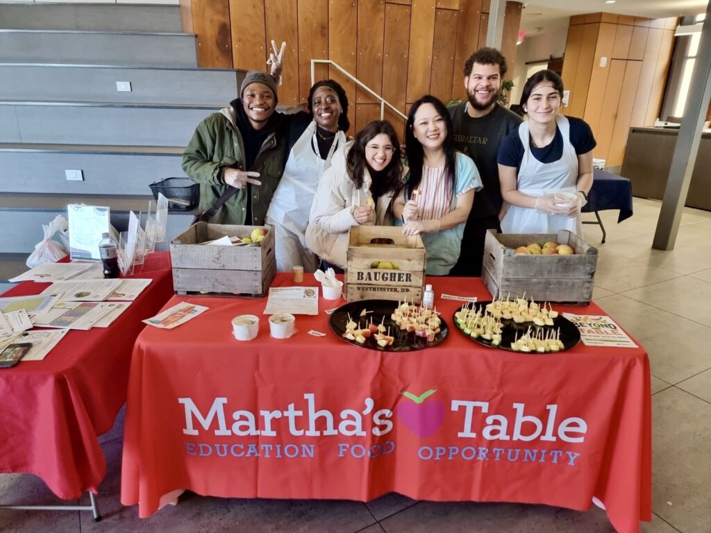 20240312_141633617_iOS Volunteers smiling behind a Martha's Table booth with fruit displays and educational flyers.