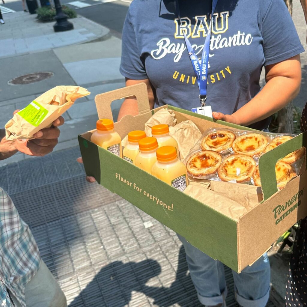 IMG_0254 Person holding a tray of pastries and juice outdoors, next to someone with a sandwich, representing college refreshment.