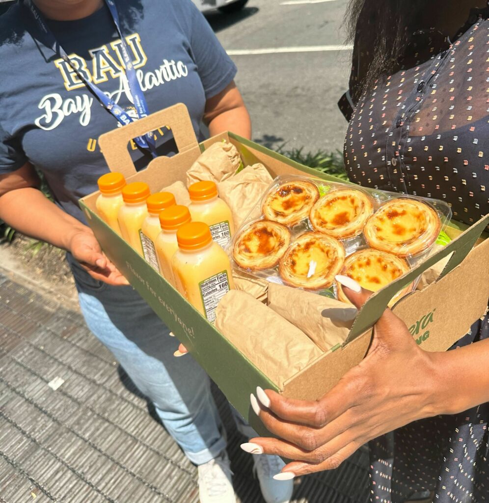 IMG_0255 Box of orange juice bottles and custard tarts being held outdoors by two people in sunny weather.