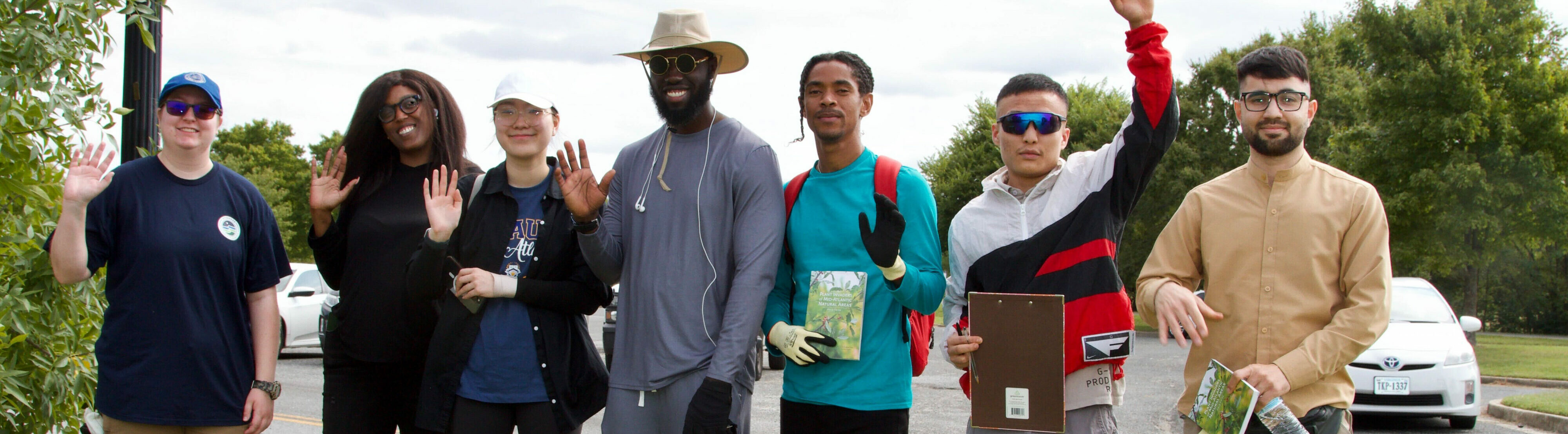 A diverse group of people smiling and waving outdoors, participating in a community event or nature walk.