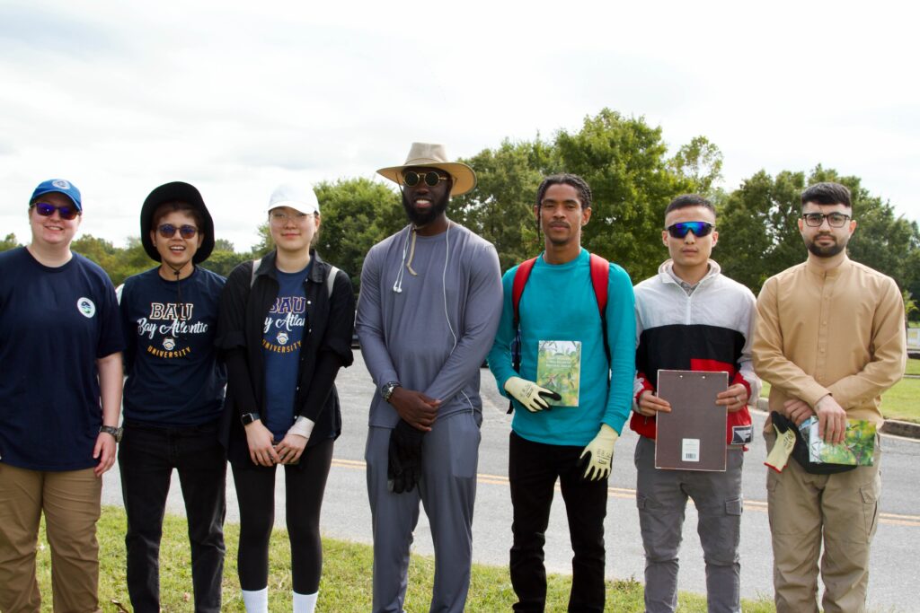 IMG_2859 A diverse group of seven students standing outdoors, holding books, and wearing varied casual attire, smiling at the camera.