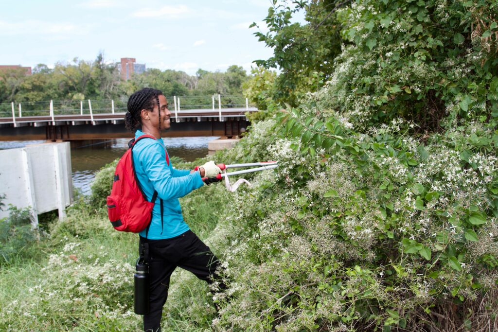 IMG_2861 Young man trims overgrown greenery by a river with a bridge in the background, wearing a red backpack and gloves.
