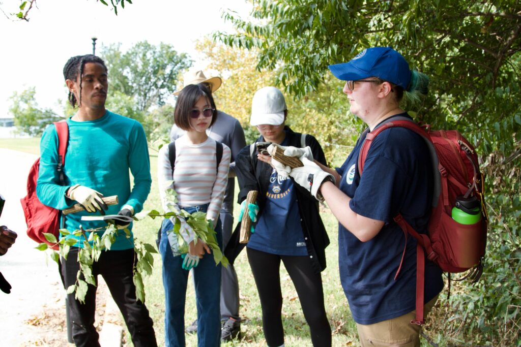 IMG_2864 Group of people learning about plants outdoors, holding branches and listening to a guide in a park setting.
