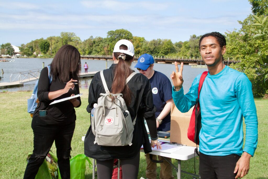 IMG_2869 People at an outdoor event by a river, some registering at a table; one person signs, another gestures a peace sign.