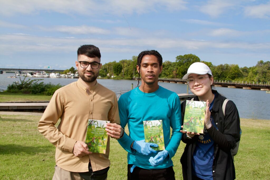 IMG_2870 Three people outdoors holding booklets on plant invasions, with a river and boats in the background.