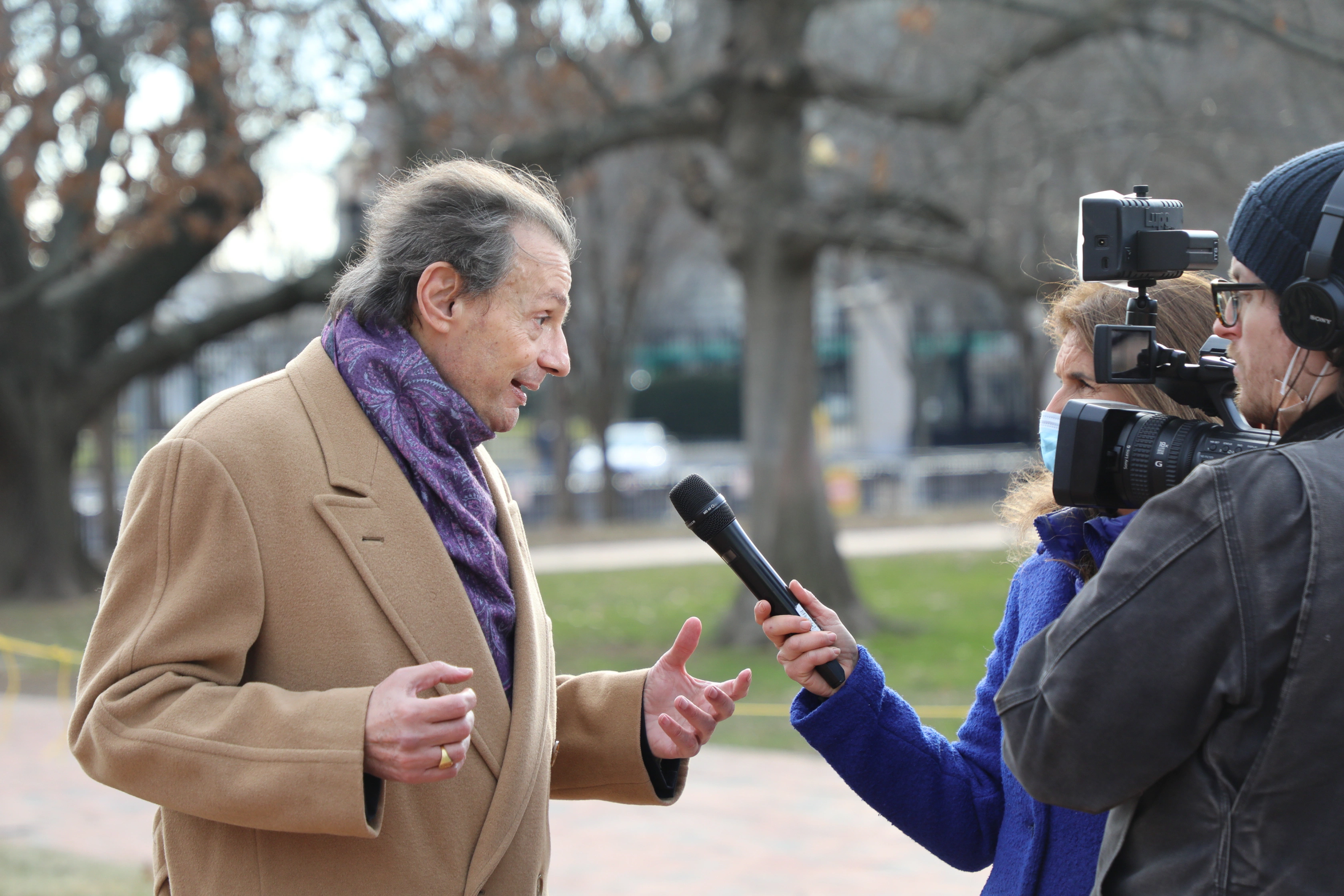 Man in coat and scarf gives interview to reporter with camera in park.