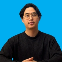 Man with glasses and a black shirt against a blue background, sitting and facing forward.