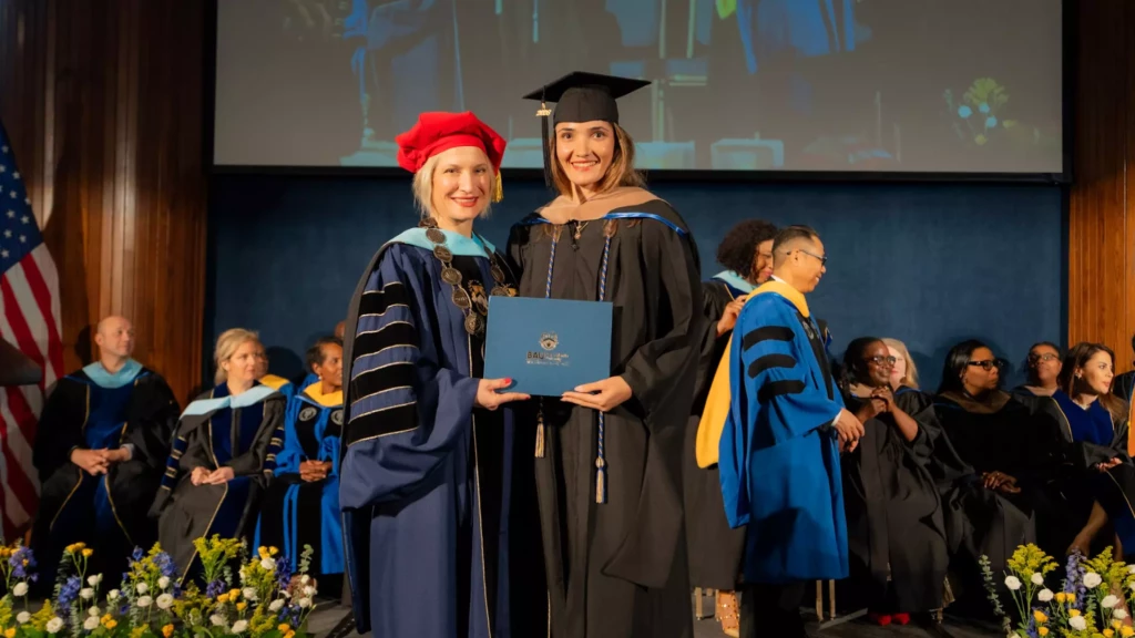 Graduate receives diploma on stage during ceremony, surrounded by faculty in academic regalia.