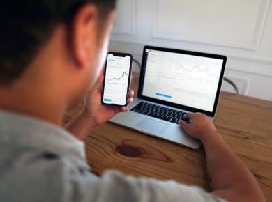 How Much Do Data Analysts Make & How to Become One (2025) 22 Man analyzing stock market graphs on phone and laptop screens at a wooden desk.