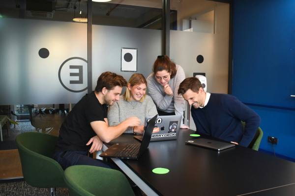 Group of young professionals collaborating and smiling around a laptop in a modern office, demonstrating communication skills in tech jobs