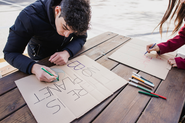 Two people creating climate change protest signs on cardboard with markers at an outdoor table.