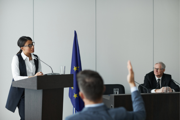Woman speaking at a podium during a conference, European flag in the background, audience member raising hand.