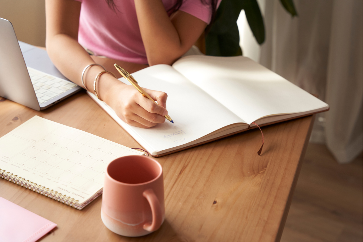 Person writing in notebook at desk with laptop, calendar, and pink mug, suggesting planning or journaling.