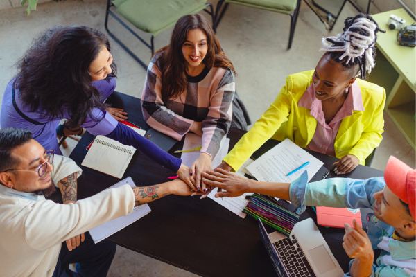 A diverse team collaborating, stacking hands over a table with notebooks and a laptop, showing unity and teamwork.