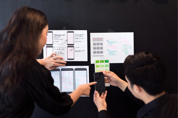 Designers collaborating on mobile app wireframes, reviewing layouts and interface prototypes on a black wall.