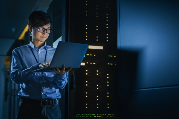 cybersecurity graduates works on a laptop in a dimly lit server room, managing network hardware and data security.
