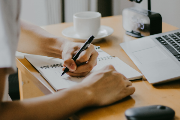 Person writing in a notebook at a desk with a laptop and coffee, suggesting productivity and focus.