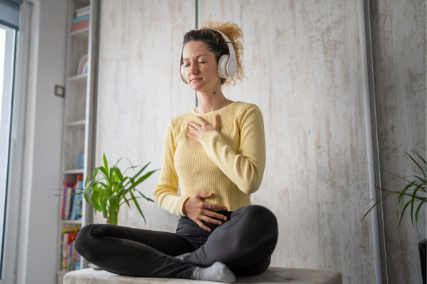 Woman meditating with headphones, sitting cross-legged indoors, eyes closed, wearing a yellow sweater, near plants.