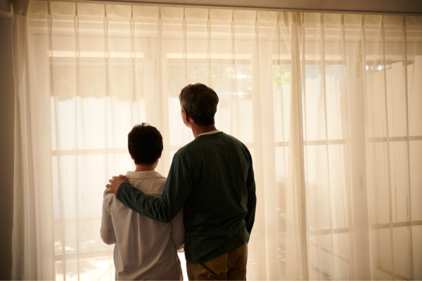 Two people standing together, one with an arm around the other, looking out of a window with light curtains.