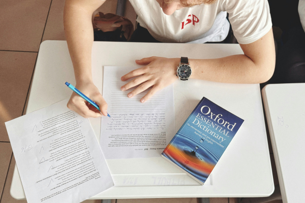 Student writing notes with a blue pen at a desk, Oxford Essential Dictionary nearby, emphasizing study and learning.