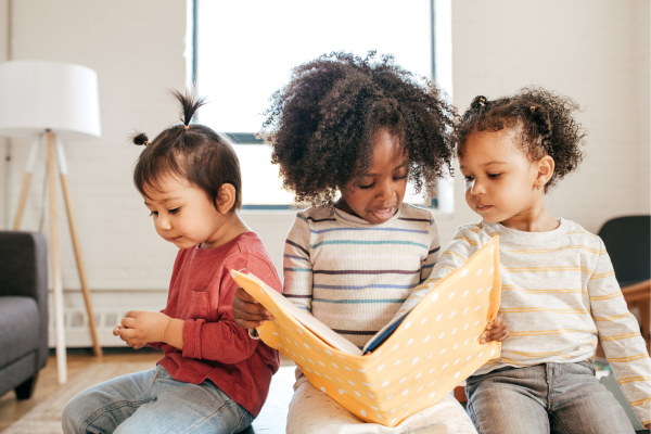 Three children sitting together, reading a large yellow book in a cozy room with natural light and a floor lamp.