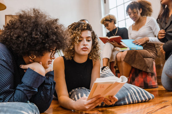 Group of young people in student clubs reading and studying together on the floor in a cozy room with wooden floors and pillows.
