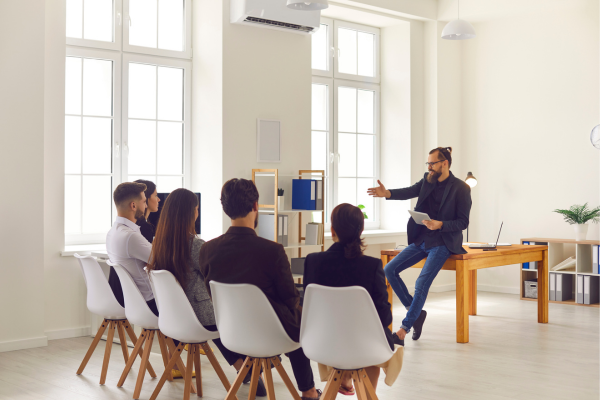 Man leads a team meeting in a modern office space, engaging with diverse colleagues seated on white chairs.