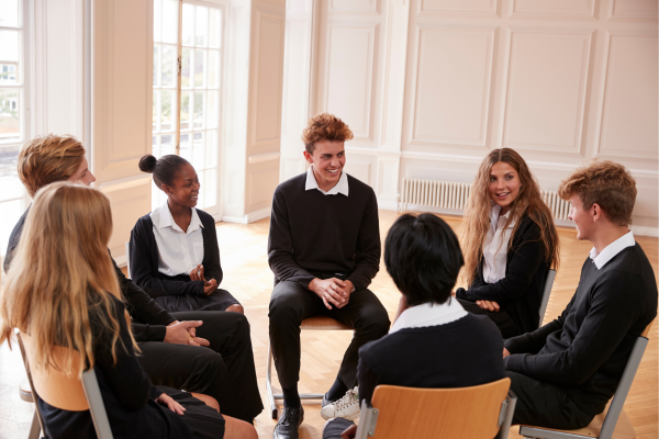 Members of student clubs in school uniforms sitting in a circle, engaged in a group discussion in a bright classroom.