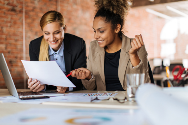 Two businesswomen discussing documents and ideas in a modern office setting, with a laptop and charts on the table.