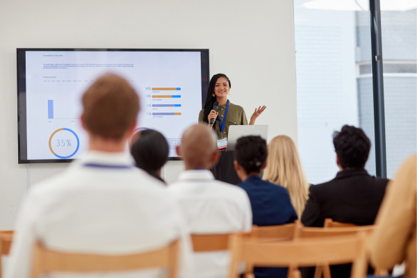 A woman giving a presentation to an audience with a screen showing charts and graphs in a conference setting.