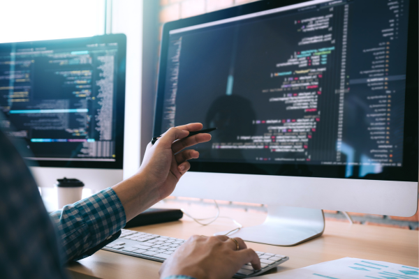 Person coding on computer with multiple screens displaying source code in a modern office setting.