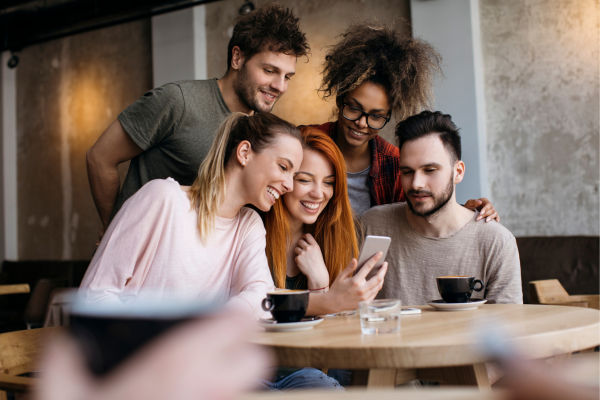 Group of friends in a cafe smiling at a smartphone together, enjoying coffee and socializing.