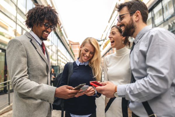 Group of four diverse professionals smiling and sharing content on smartphones outside modern office building.