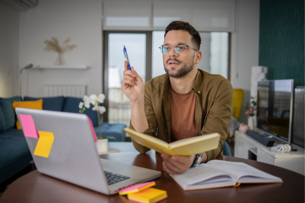 Man working from home, brainstorming ideas while holding a pen and taking notes on a laptop and notebook.