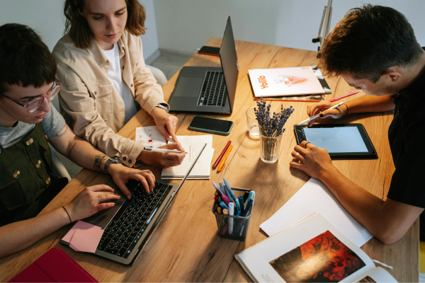The Rise of Social Entrepreneurship: Combining Business with Purpose 9 Three people collaborating at a wooden desk with laptops, notebooks, and digital tablets for a creative project.