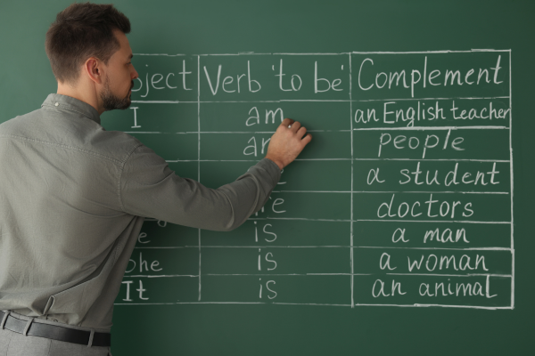 10 Essential Tips for New ESL Teachers: How to Teach English Like a Pro 8 Man teaching English grammar on chalkboard, writing verb to be and complements in classroom setting.