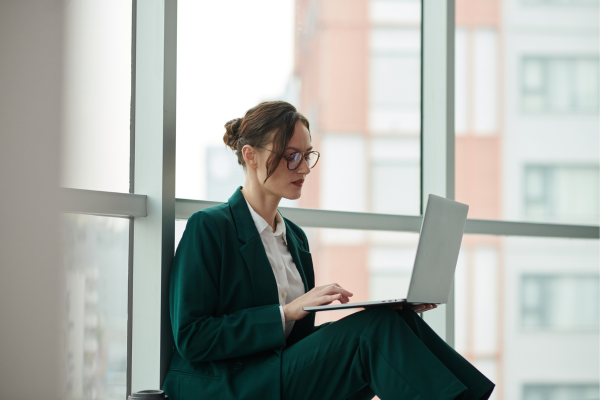 Business Analyst Jobs in Global Demand: Skills, Careers, and Opportunities 5 A woman in a green suit works on a laptop while sitting on a windowsill in an office.