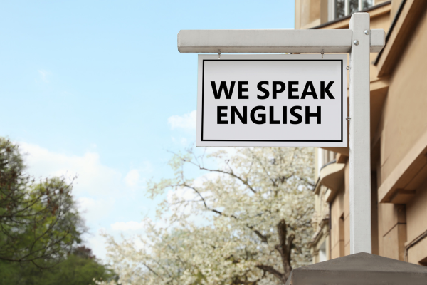 TOEFL vs. IELTS: Which English Proficiency Test Should You Take? 6 Sign on a building with We Speak English message, set against a background of trees and a clear sky.