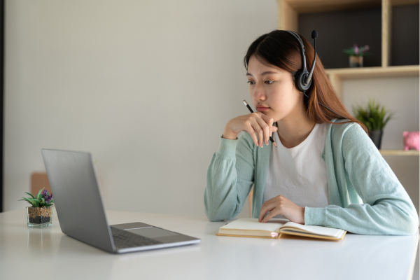 TOEFL vs. IELTS: Which English Proficiency Test Should You Take? 7 Woman wearing headset studying online at home with laptop and notebook.